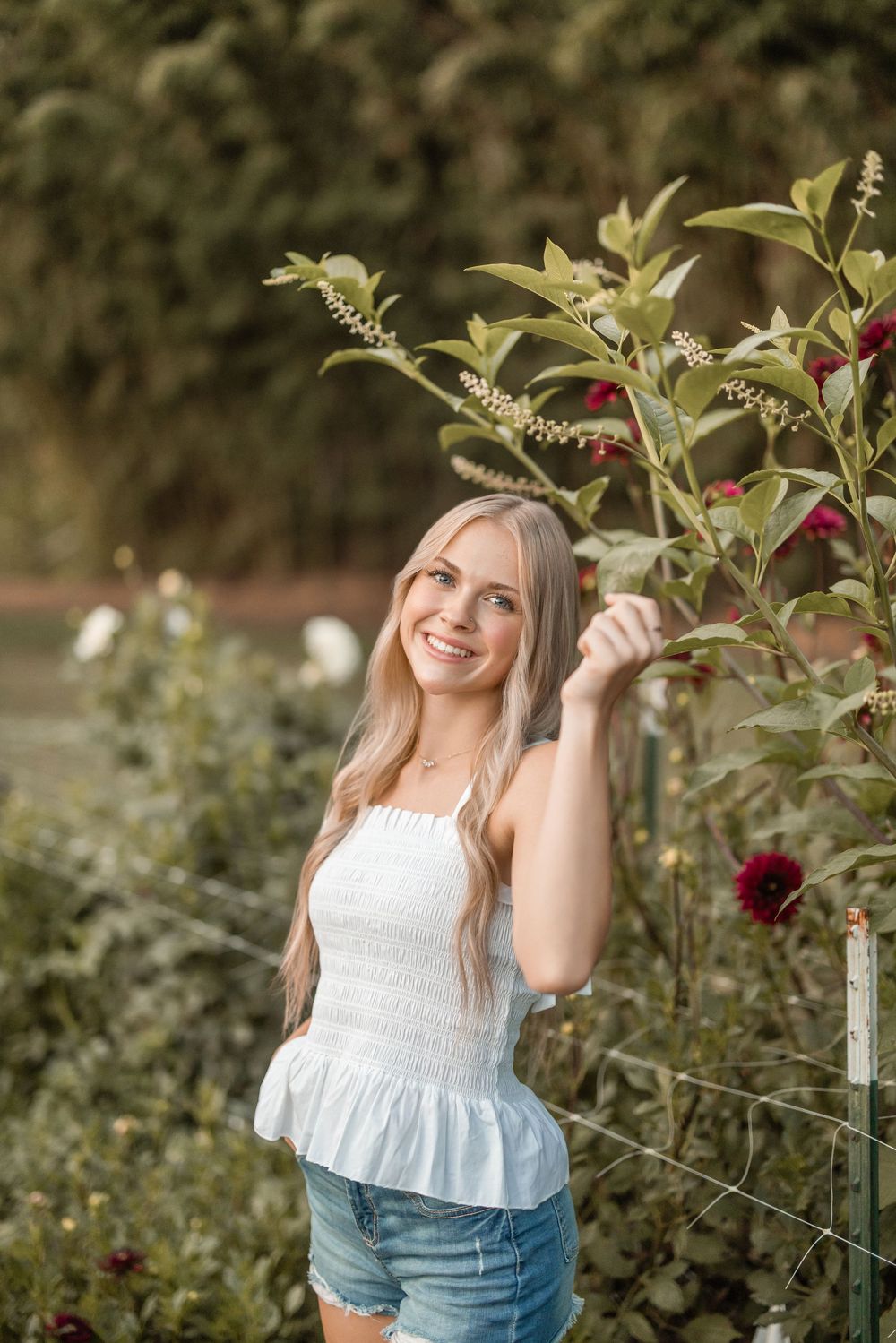 A person in a white top and denim shorts poses in a garden next to blooming red flowers and greenery.