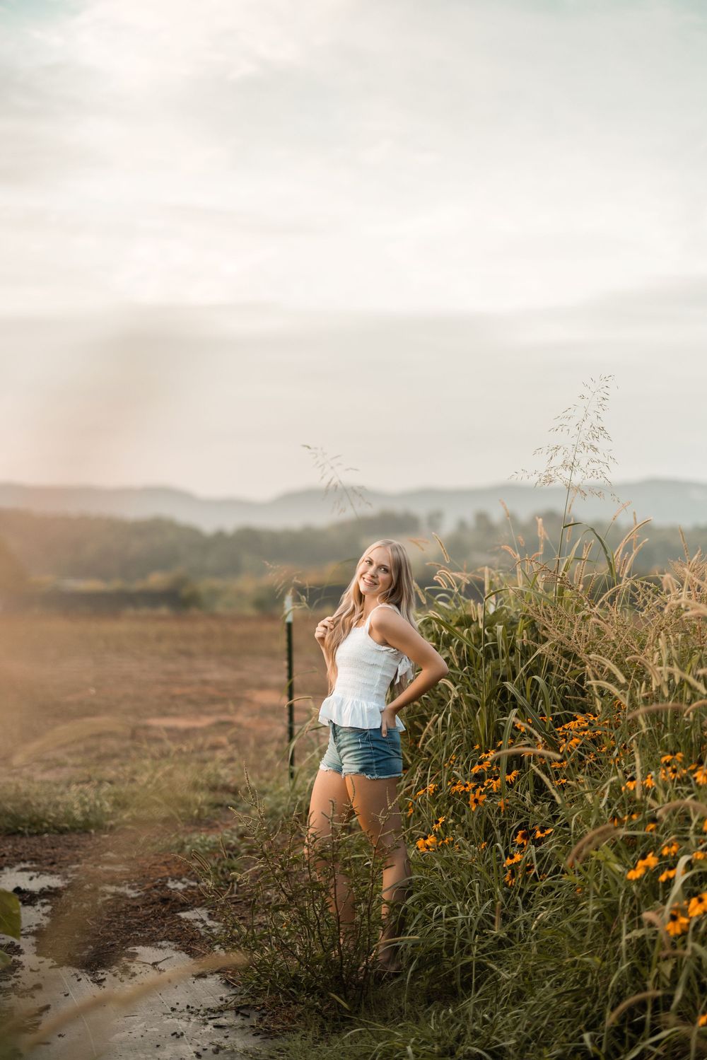 A woman stands amidst tall wildflowers on a misty hillside, wearing denim shorts and a white tank top.