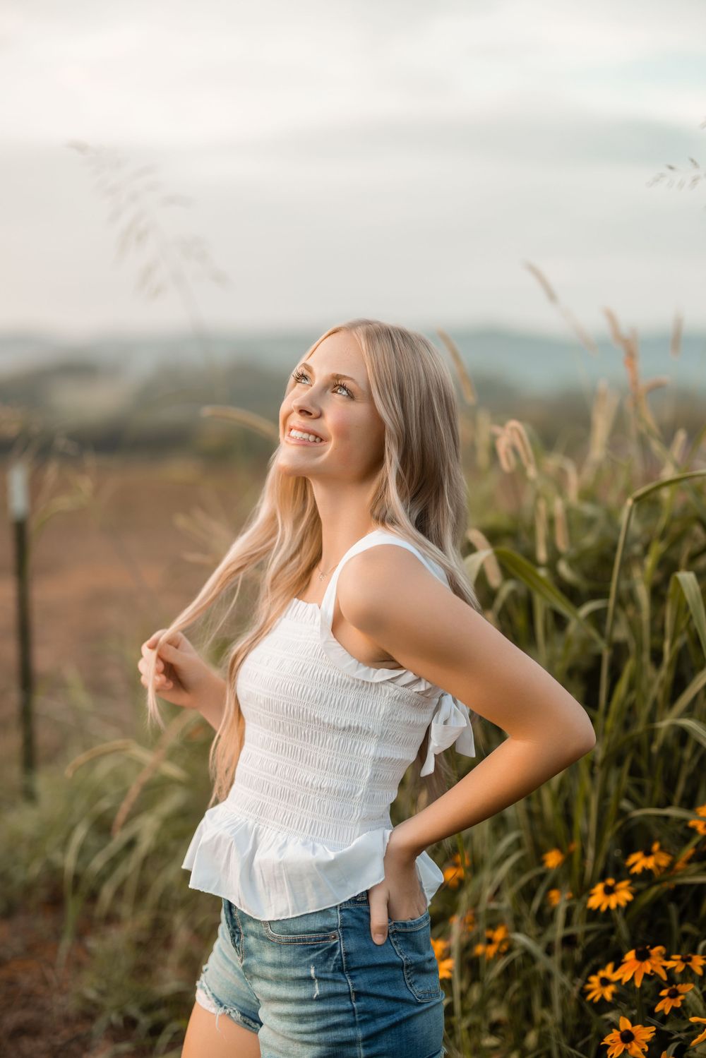 Person in white sleeveless top and jeans poses outdoors among wildflowers at golden hour.