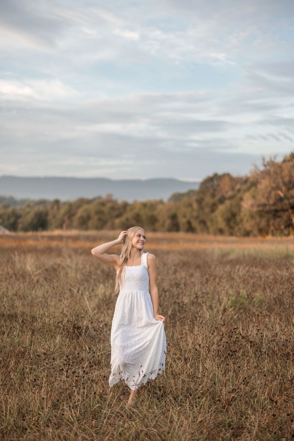 A flowing white sundress twirls in a golden autumn field with mountains and trees in the distance at sunset.
