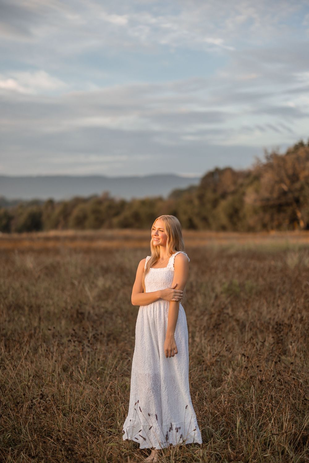 A figure in a flowing white sundress stands in a golden field at sunset with mountains in the distance.