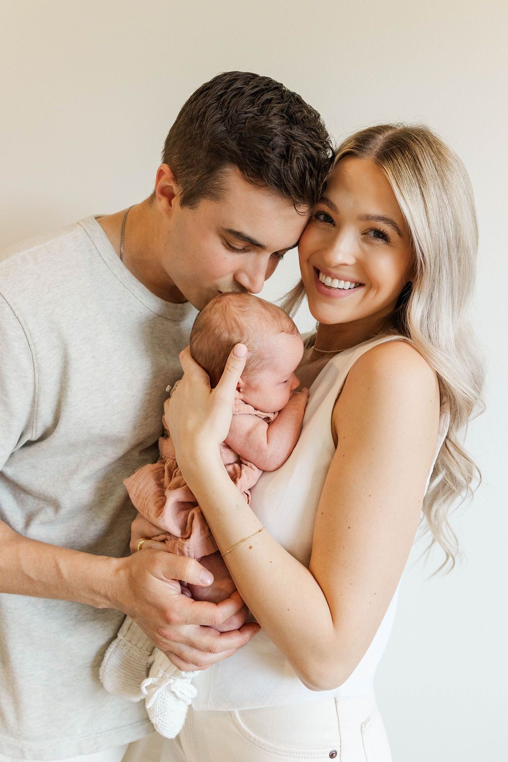 Happy new parents smiling and kissing their newborn baby wrapped in pink blanket indoors.