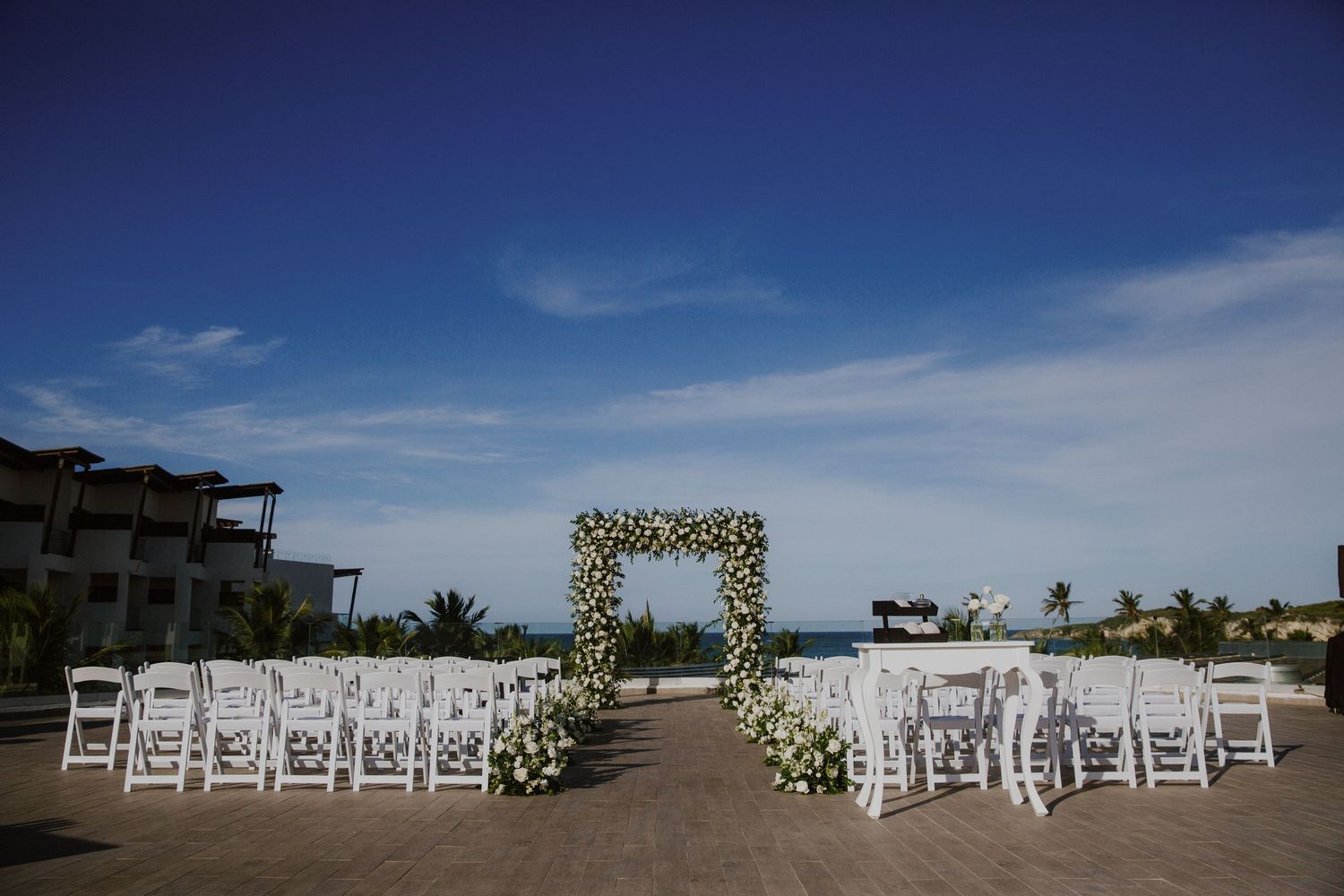 Outdoor wedding ceremony setup with white floral arch against blue sky and ocean backdrop.