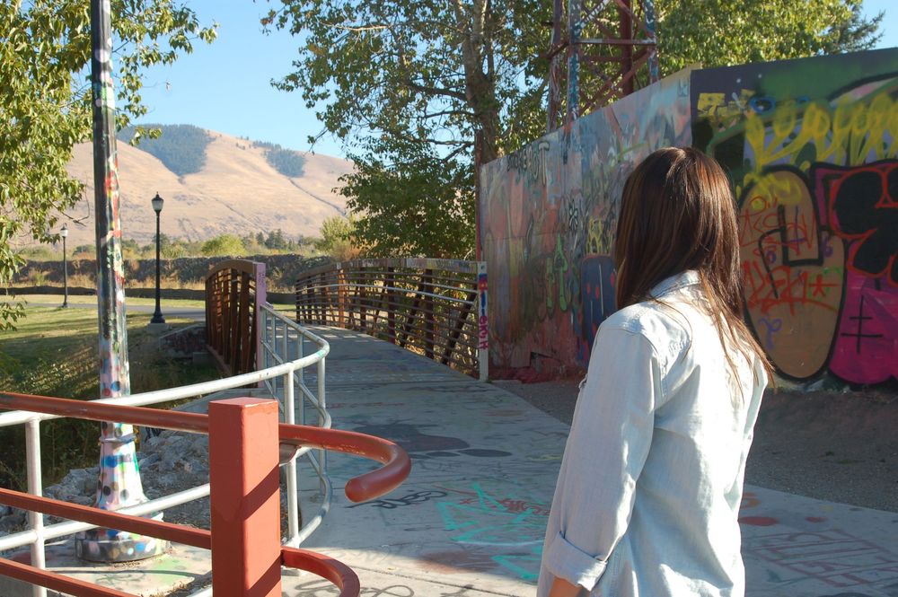 A young woman with brown hair, seen from behind, is standing on a concrete path next to a fence with red railing. She is wearing a light bl
