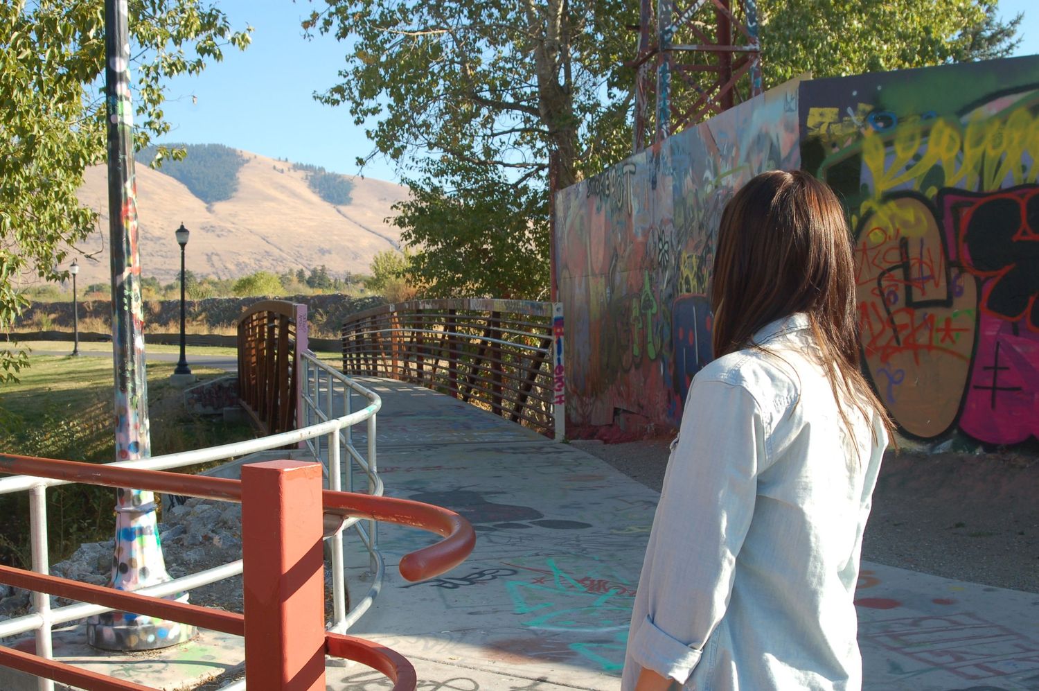 A young woman with brown hair, seen from behind, is standing on a concrete path next to a fence with red railing. She is wearing a light bl