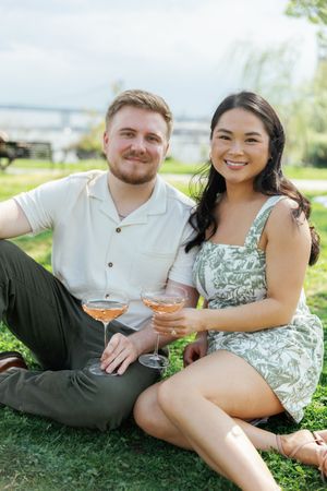 Couple enjoys cocktails while sitting on grass during outdoor summer picnic near waterfront.