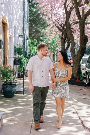 A couple strolls together under blooming cherry blossom trees along a sunlit sidewalk during springtime.