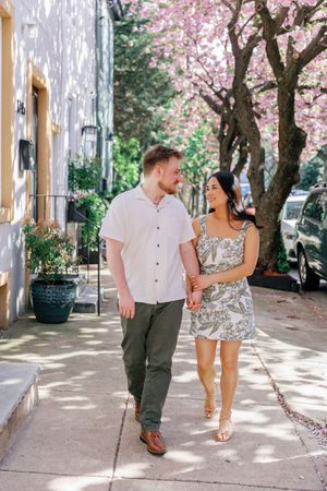 A couple walks together on a charming city sidewalk beneath flowering pink cherry blossoms on a sunny spring day.