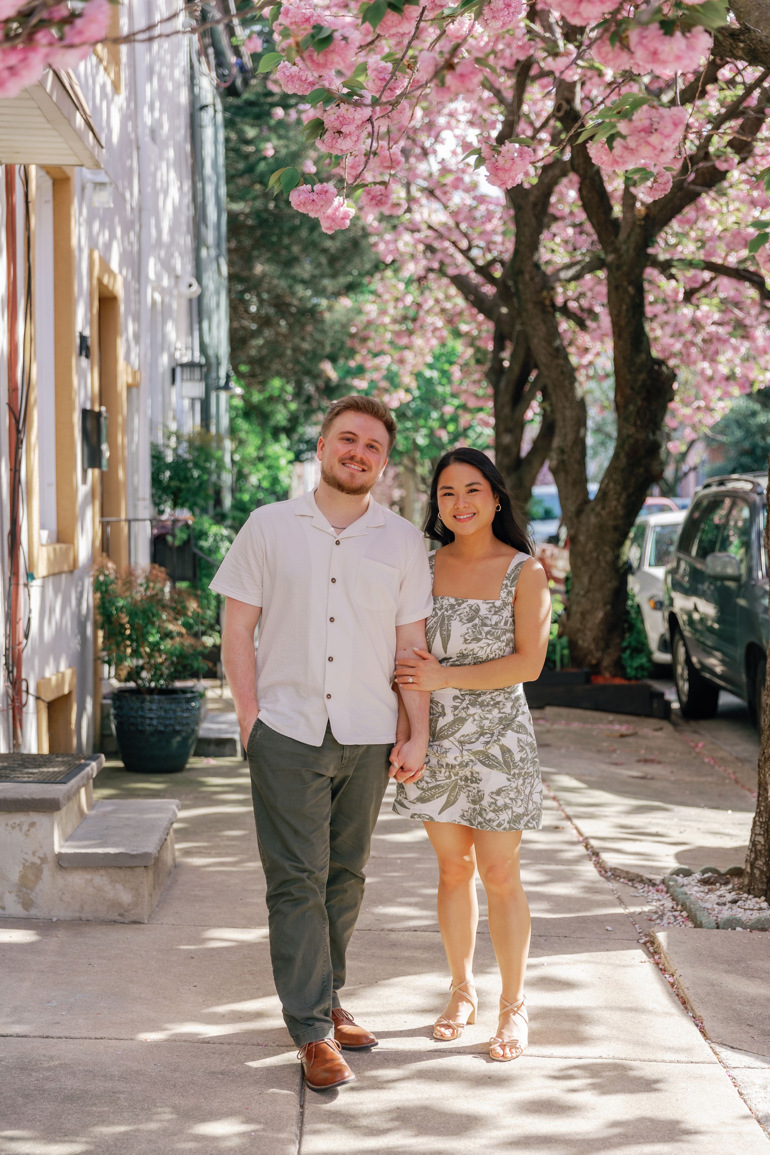 Couple walking hand in hand under blooming pink cherry blossom trees along a sunlit pathway.
