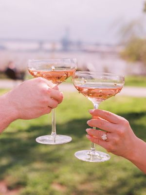 Two people toasting with champagne coupe glasses filled with rose wine in a sunny outdoor setting.