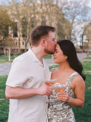 Romantic couple sharing intimate moment in park while holding cocktail glasses at sunset.