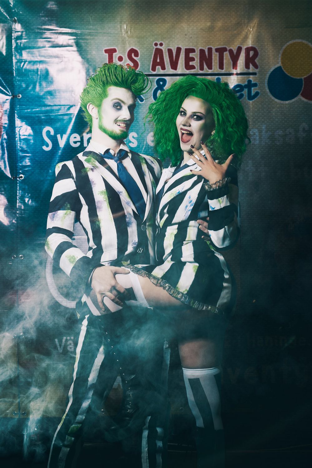 Costumed performers in striped outfits pose amid atmospheric fog with teal lighting and theatrical backdrop.