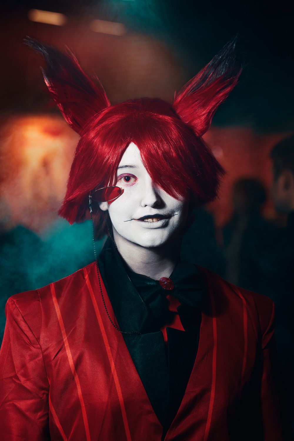 Cosplay character wearing bright red hair styled with pointed ears, a red blazer, and theatrical face paint in moody lighting.