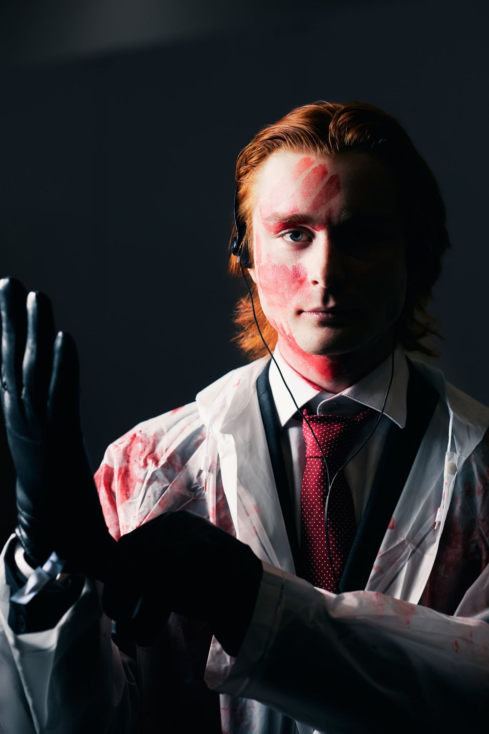 A person in a white lab coat and red tie holds up a hand in a dark moody setting with dramatic lighting.