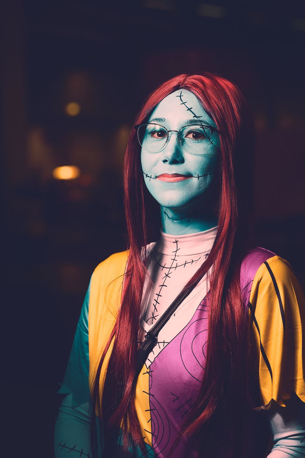 A cosplay of Sally from Nightmare Before Christmas with red hair and stitched makeup wearing colorful clothing.