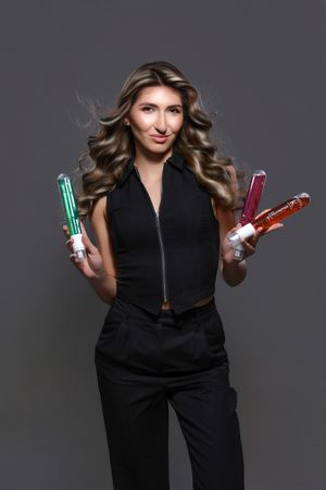 Model in black outfit holding colorful hair styling products against grey background.