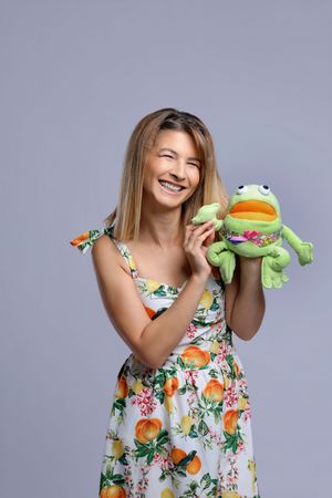 Woman in floral dress playfully poses with green frog plush toy figure against gray background.