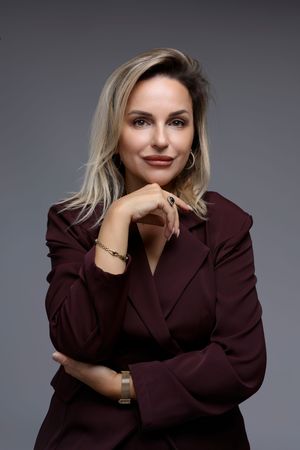 Professional woman in burgundy blazer posing confidently with hand on chin against gray background.