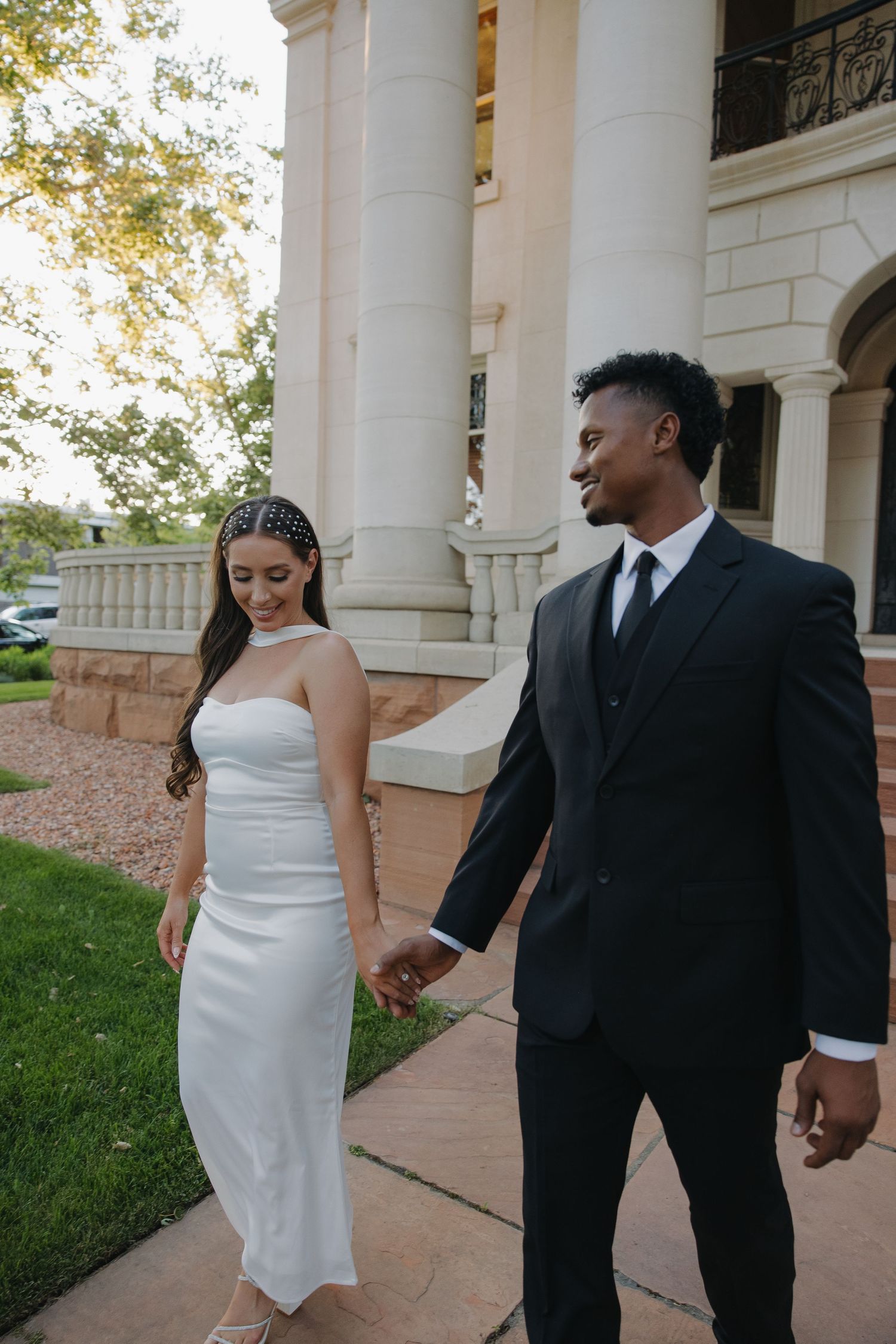 Interracial couple in wedding attire walking hand in hand outside a white columned building.