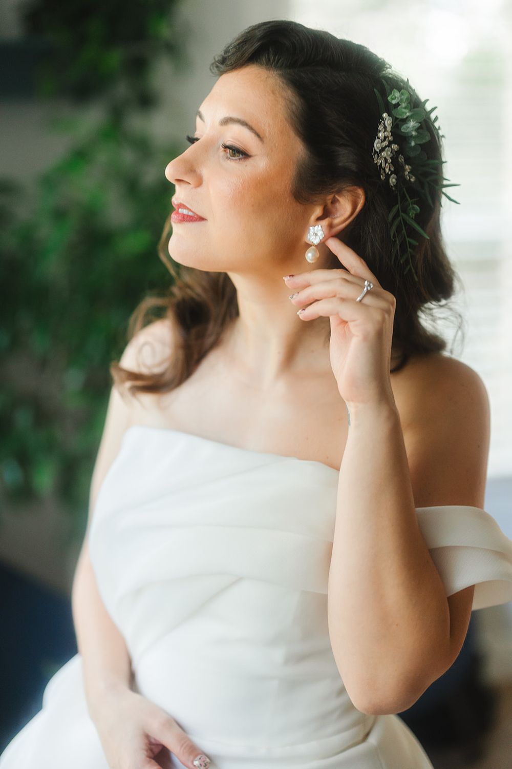 A bride in an elegant off-shoulder white gown touches her delicate earring while wearing a crystal hair accessory.