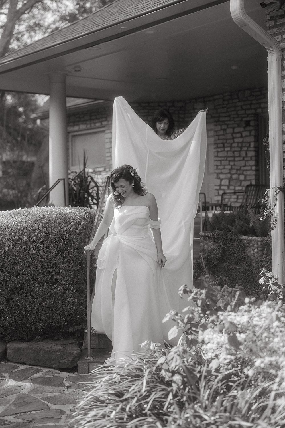 A dreamy black and white photograph shows a person in flowing white fabric standing on a garden porch with flowers.