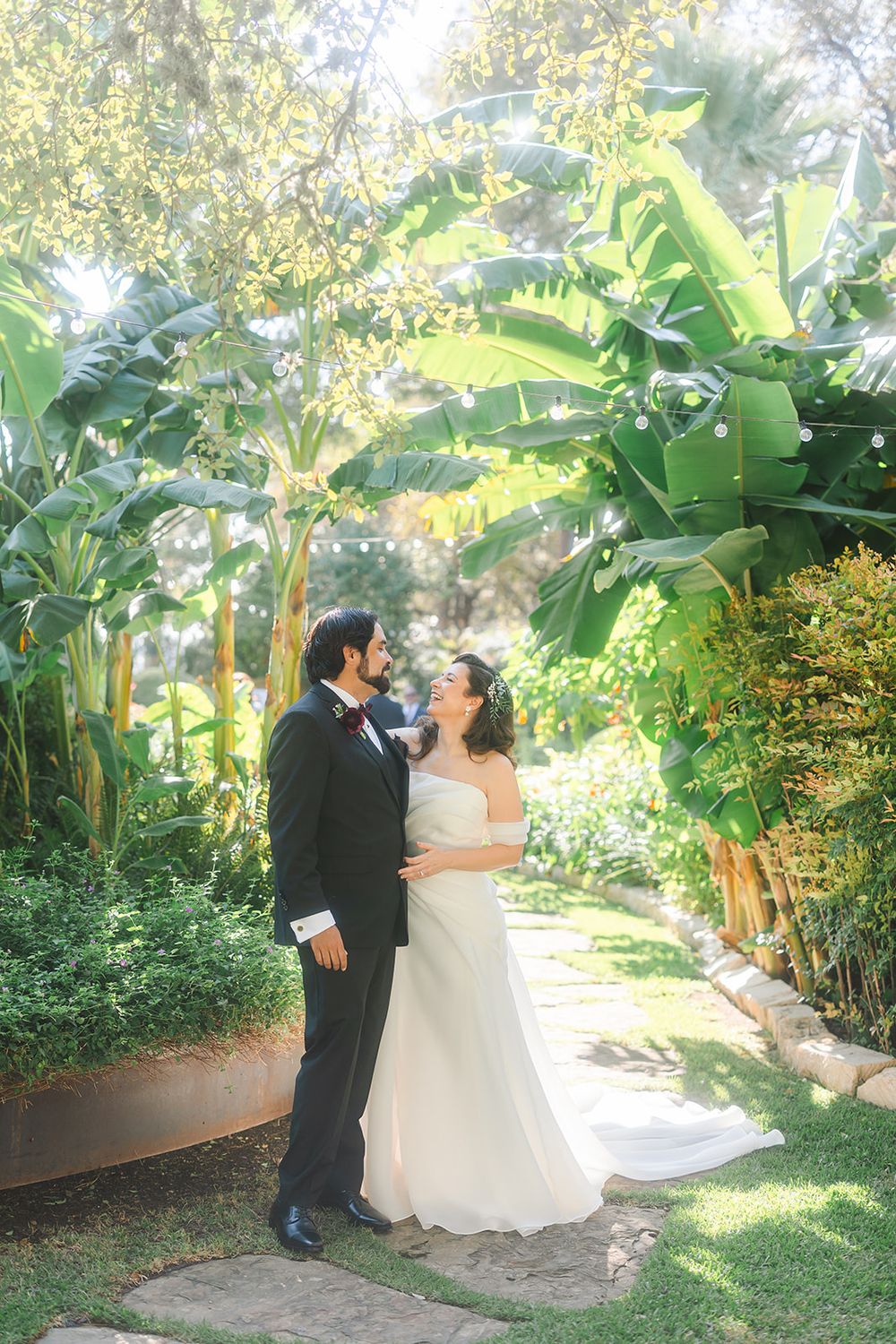A romantic wedding moment captured in a lush tropical garden with banana plants and greenery surrounding a stone pathway.