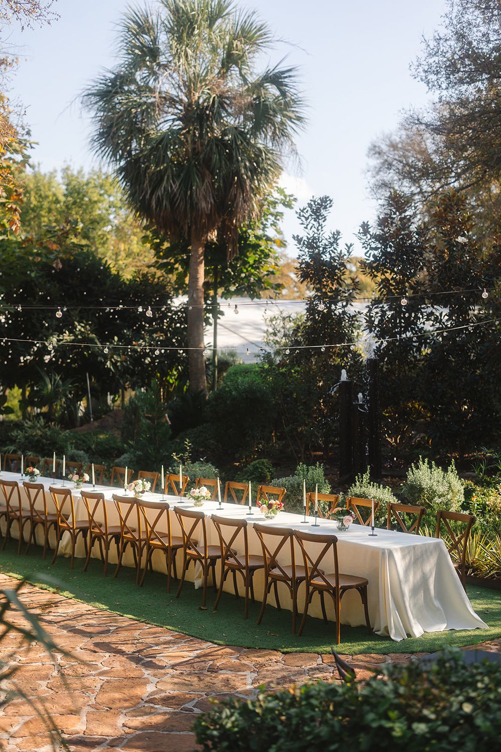 Long banquet tables with wooden chairs set up outdoors beneath palm trees for an elegant garden wedding reception.