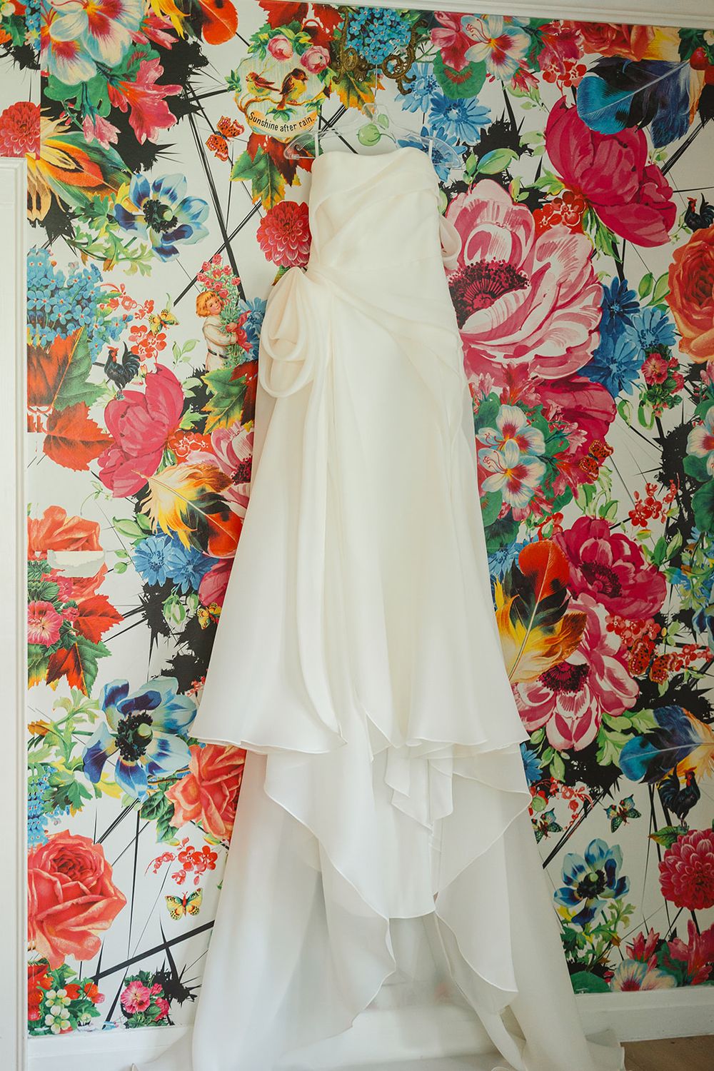 Elegant white wedding dress hanging in front of vibrant floral wallpaper with red, pink and blue blooms.