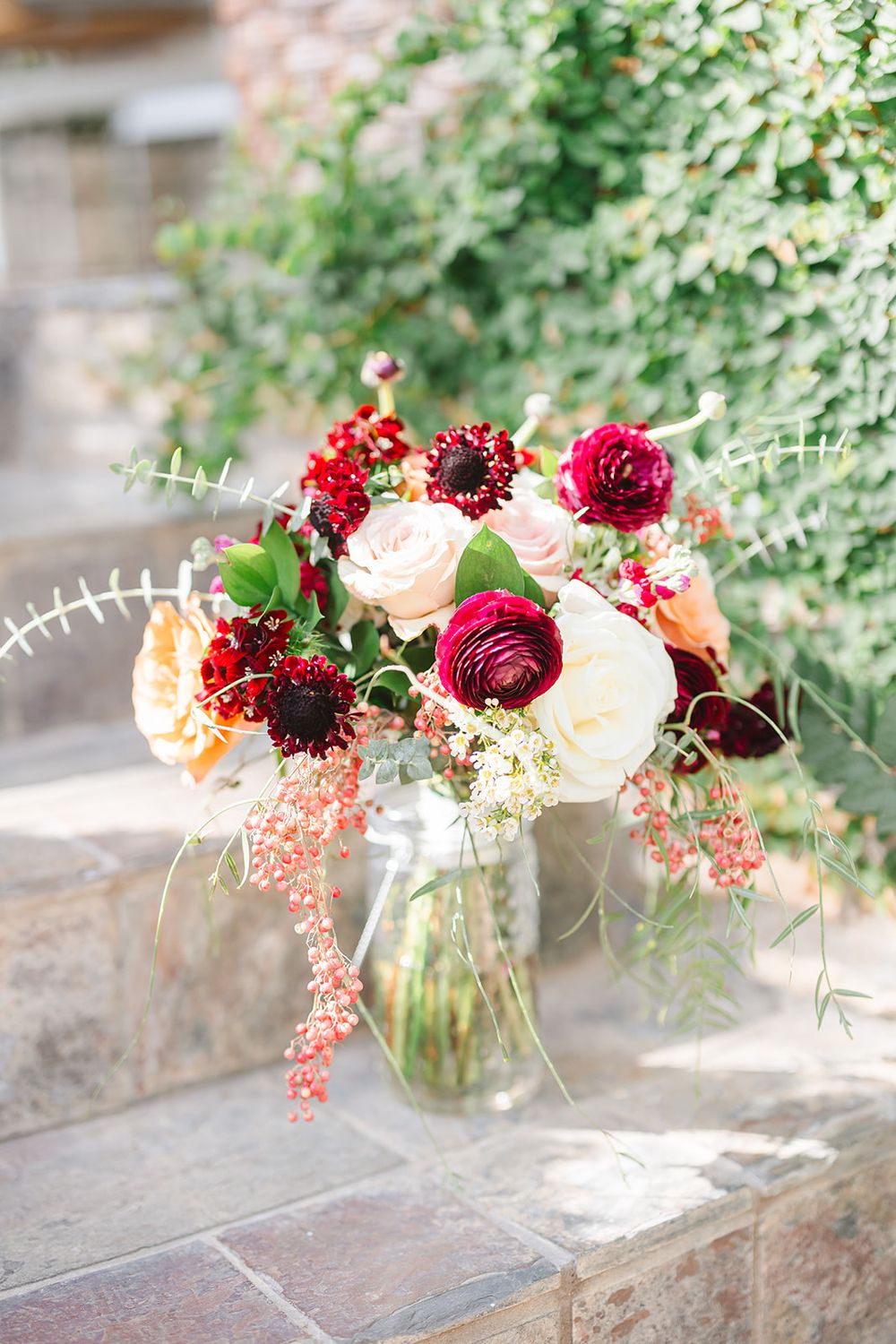 A romantic bouquet of burgundy and blush roses with cascading greenery arranged in a glass vase on stone steps.