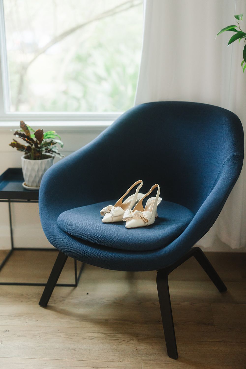 Elegant ivory bridal shoes rest on a modern navy blue accent chair near a bright window with a small potted plant.