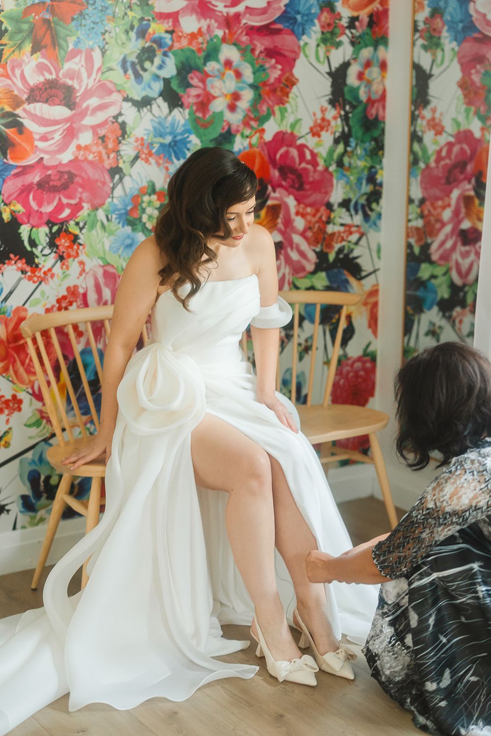 A bride in a flowing white dress sits in a chair against a vibrant floral wallpaper backdrop while getting ready.