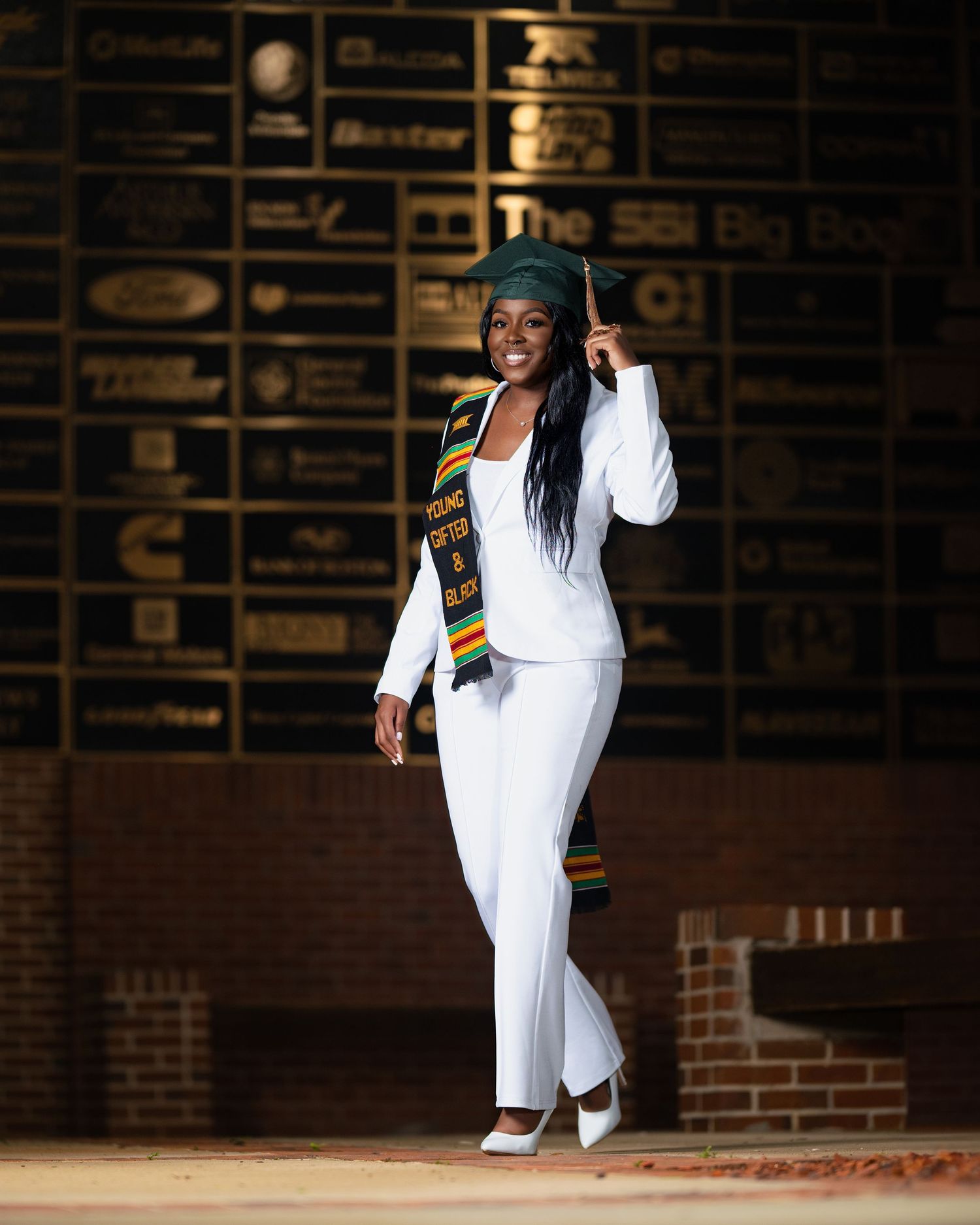 Person in white pants and shirt with African-inspired sash dances gracefully against dark brick wall backdrop.