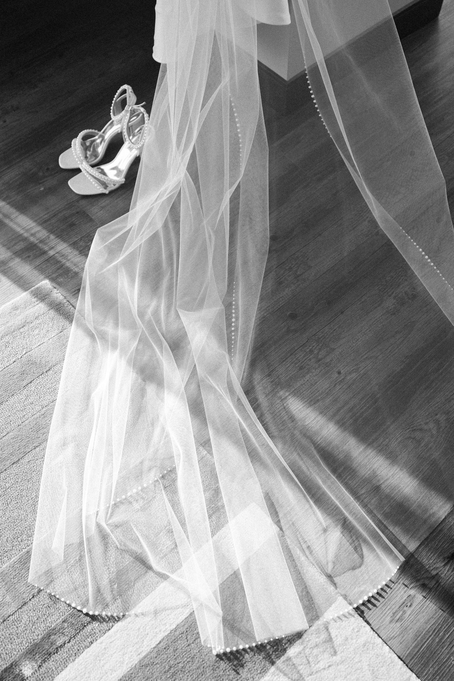Flowing white wedding dress and veil details photographed in dramatic black and white against a wooden floor.