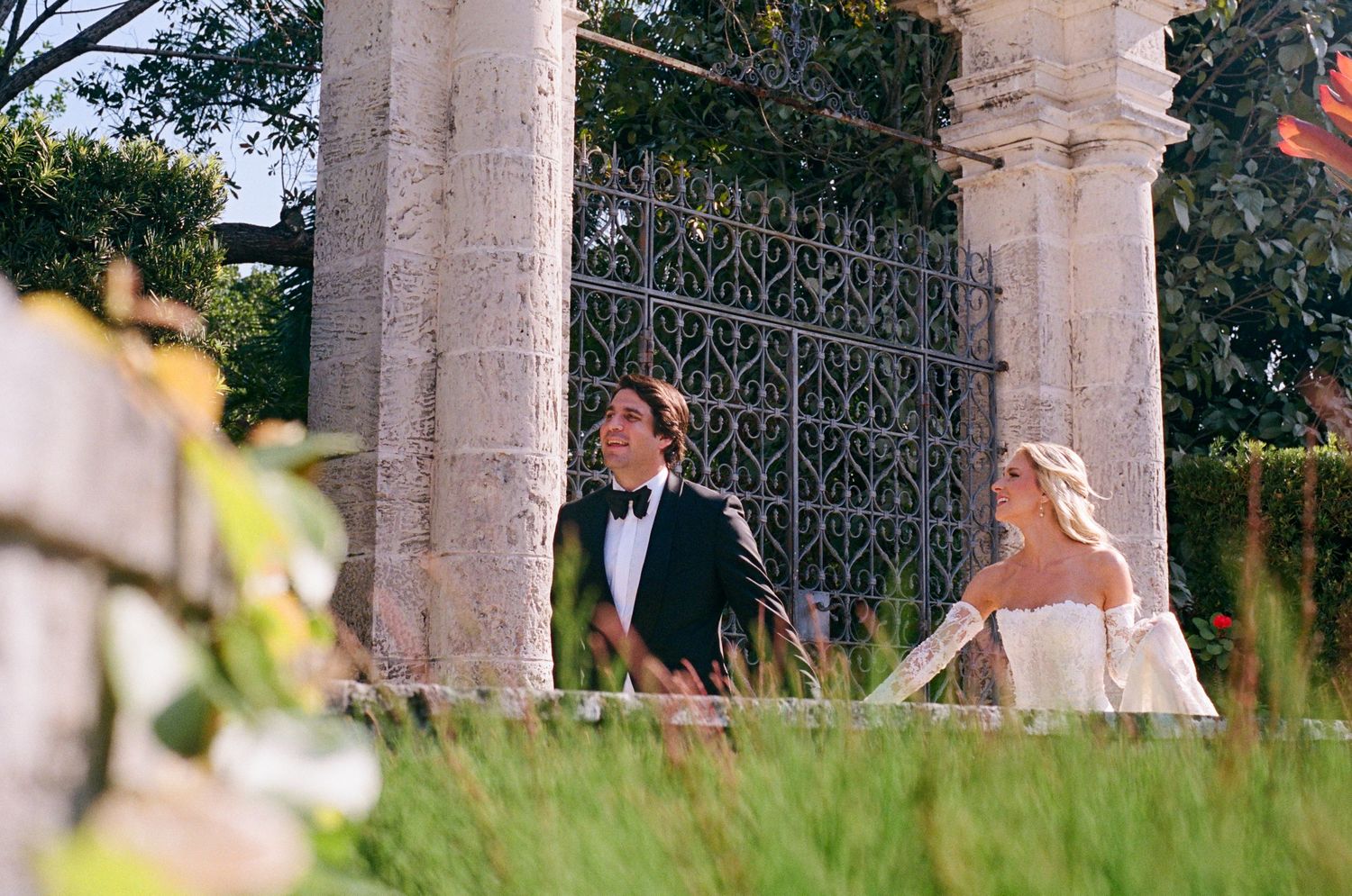A romantic wedding couple poses in an elegant courtyard with classical stone columns and decorative ironwork.