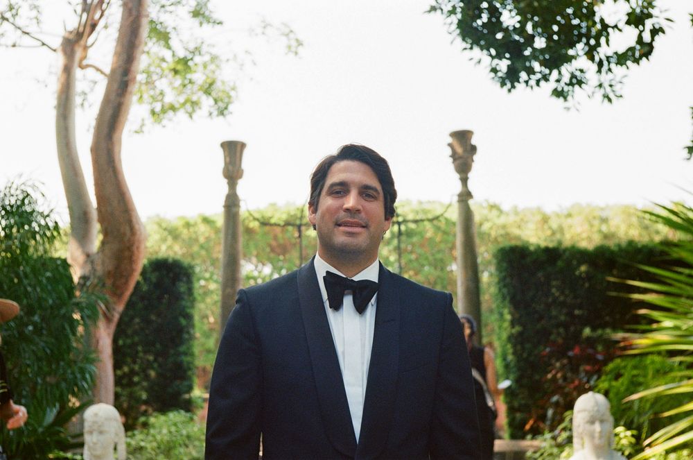 A formally dressed person in a tuxedo and bowtie stands between classical stone pillars in an elegant garden setting.