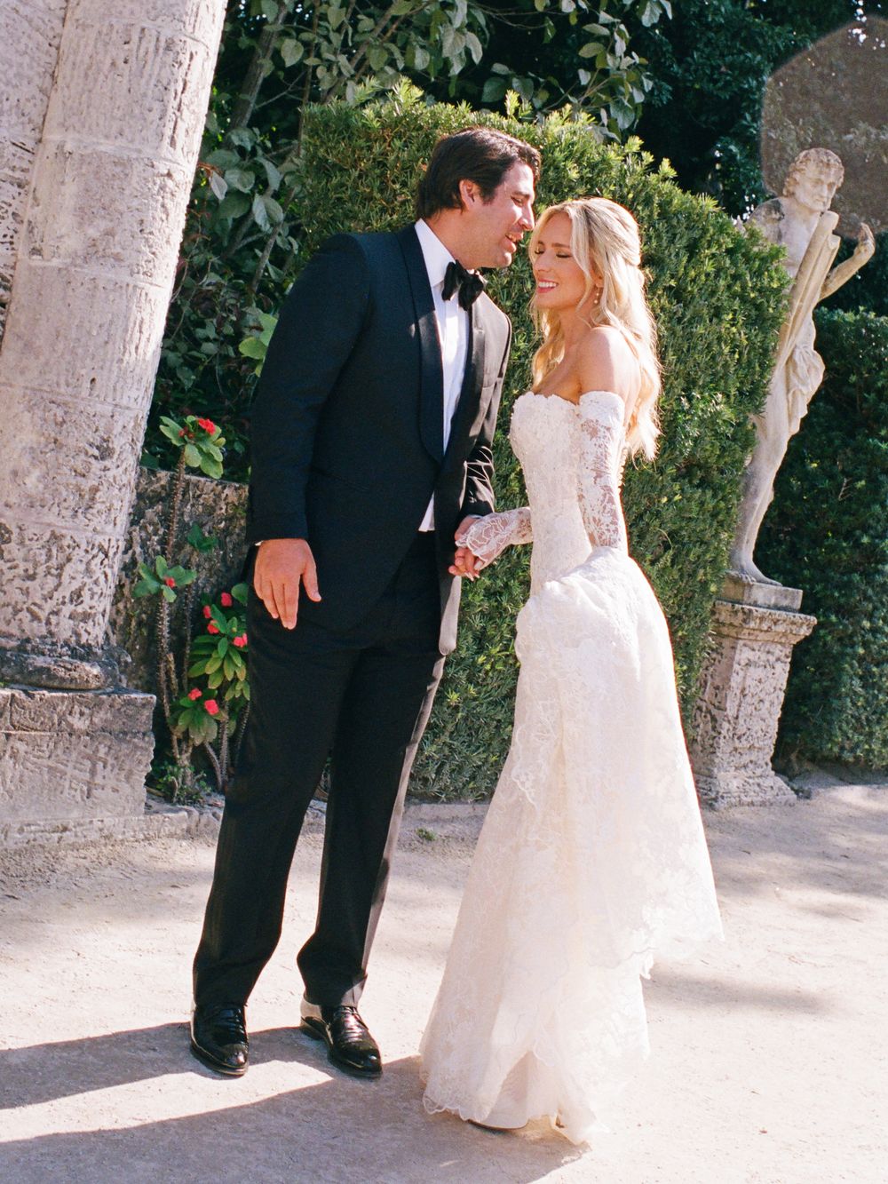 A bride in a strapless lace wedding gown shares an intimate moment with her groom in a black tuxedo outdoors.