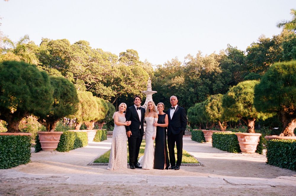 A small outdoor wedding ceremony takes place in a manicured garden with potted plants and trees at sunset.