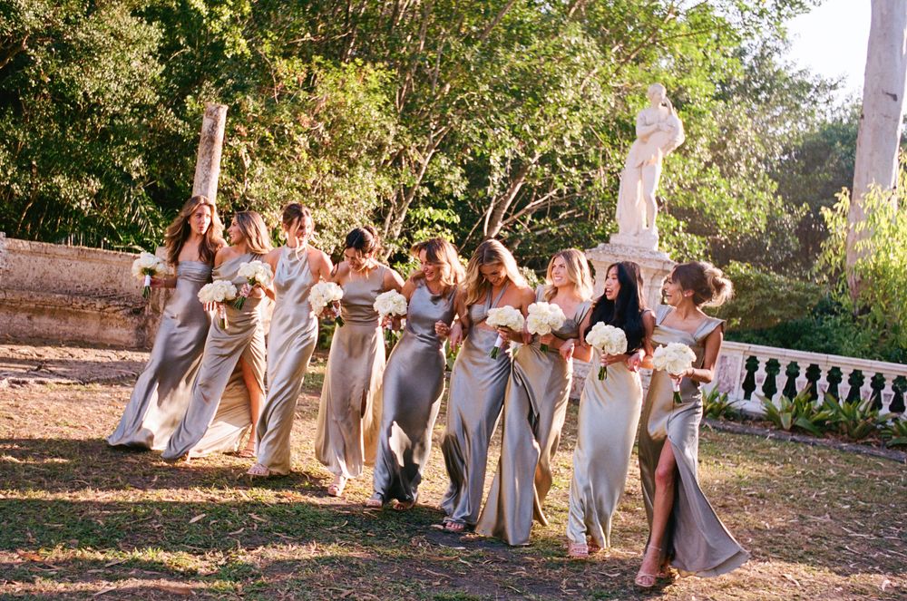 A wedding party in pale colored dresses poses together in a sunlit garden with stone balustrades.