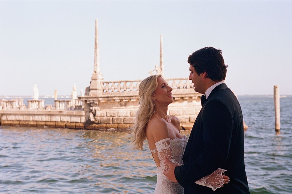 A couple standing together by a waterfront pier at sunset with ornate spires in the background.