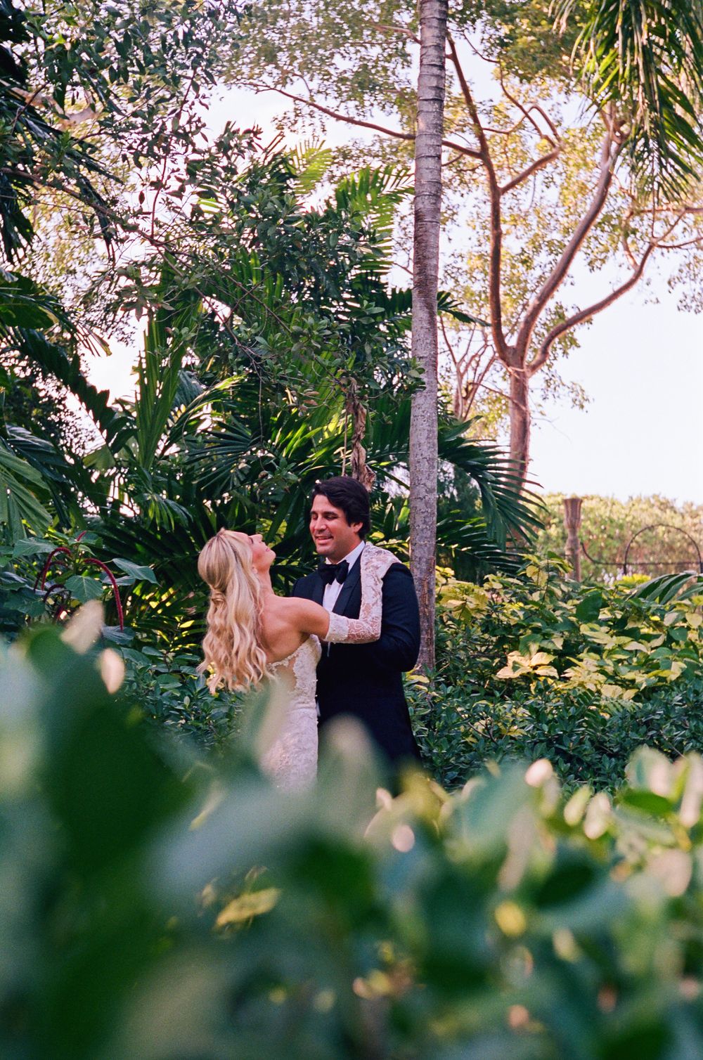 A couple shares a romantic moment surrounded by lush tropical foliage and palm trees in a garden setting.