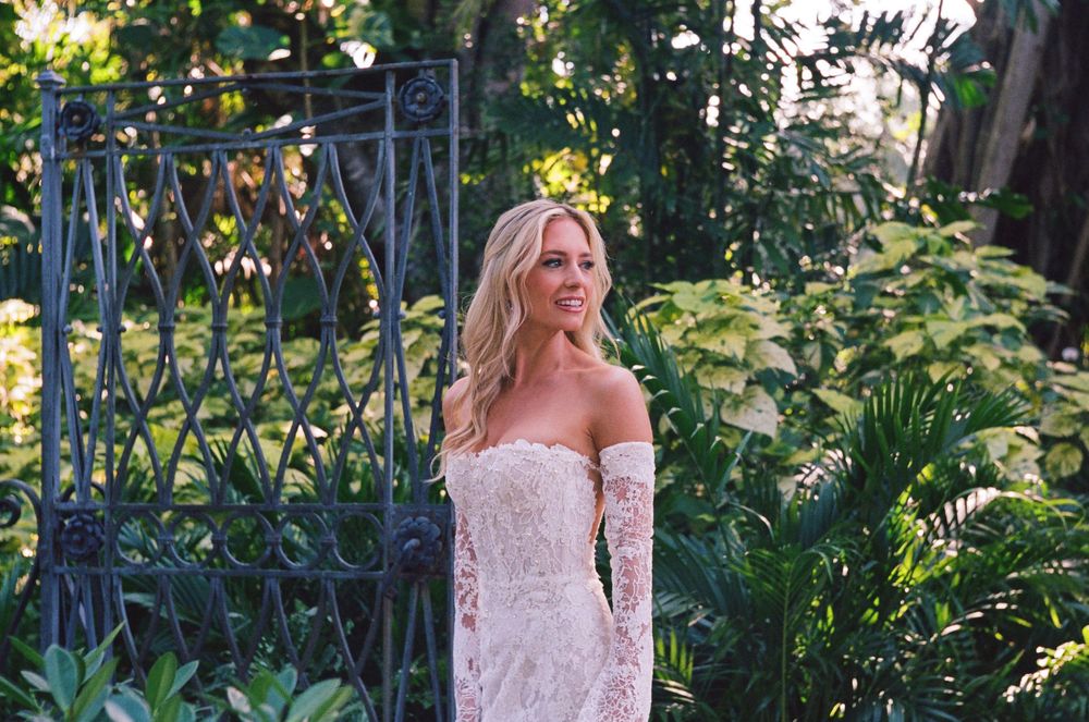 An off-shoulder lace wedding dress is shown in a lush garden setting with decorative iron gates and greenery.