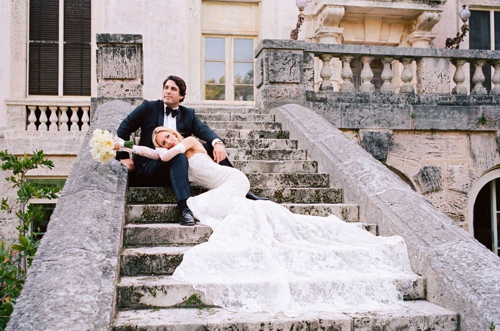 Bride in flowing white gown poses dramatically across elegant stone steps at historic European estate.