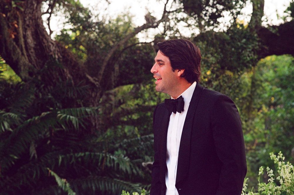 A person in a black tuxedo and bow tie stands in a lush garden setting during an outdoor formal event.