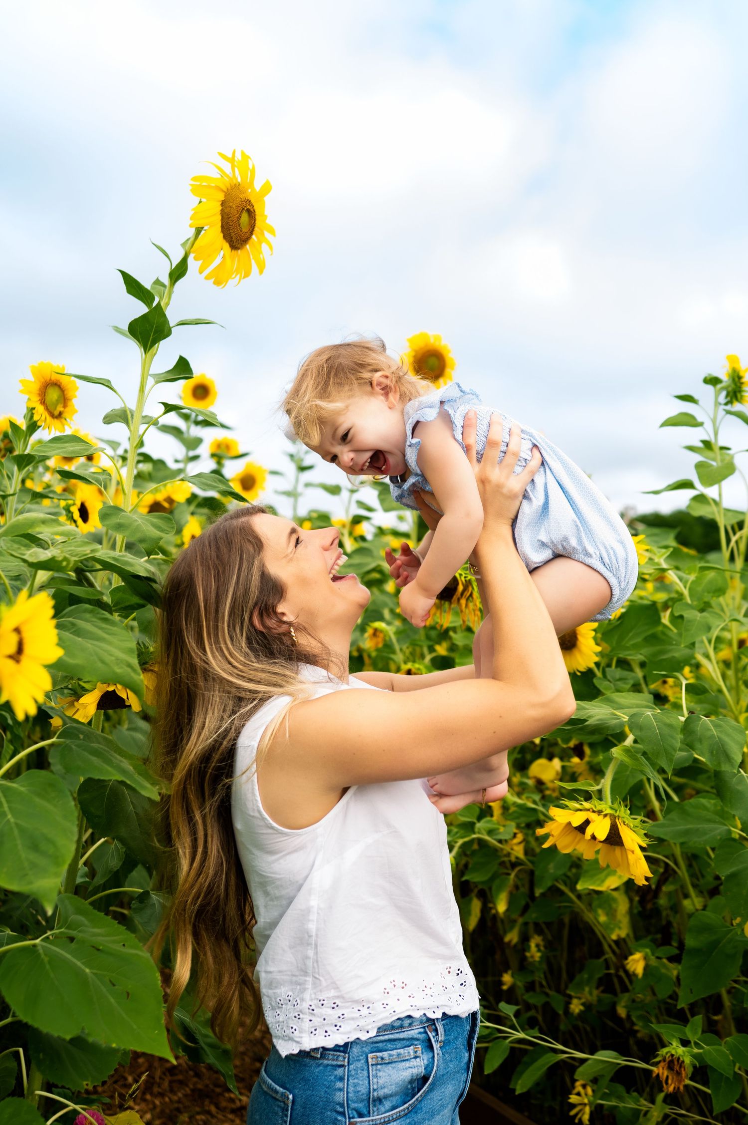 dallas mommy and me summer sunflower mini photo session; mother holds 18 month old up in a field of sunflowers under a cloudy sky photoshoot