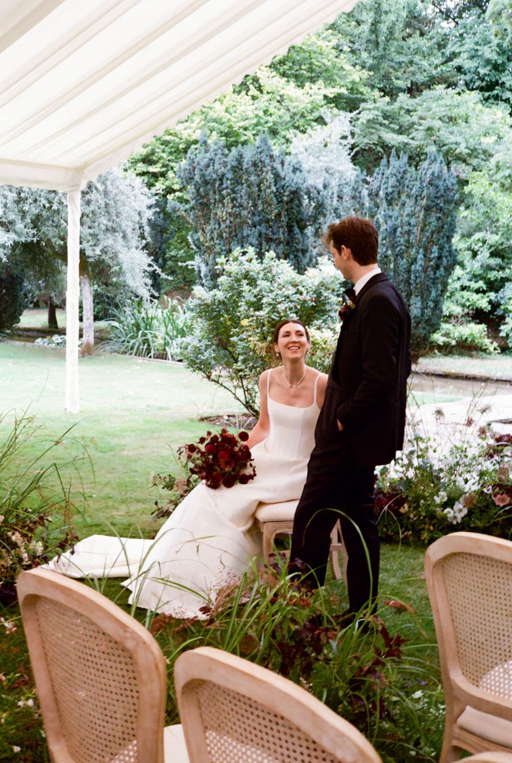 Bride & groom interacting under wedding ceremony tent. Bride seated whilst groom stands.