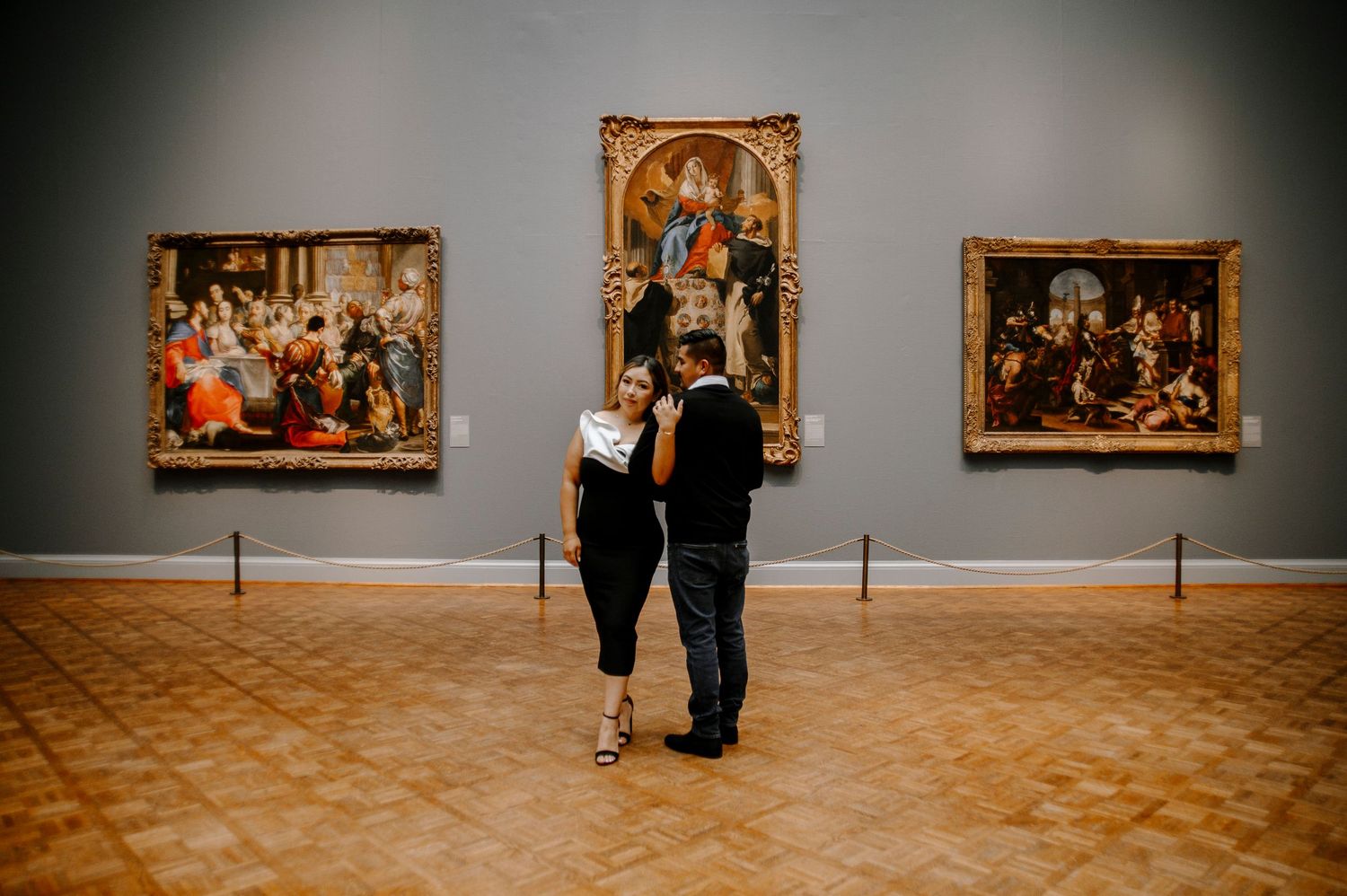 Two people walk through an art gallery with paintings displayed on gray walls.