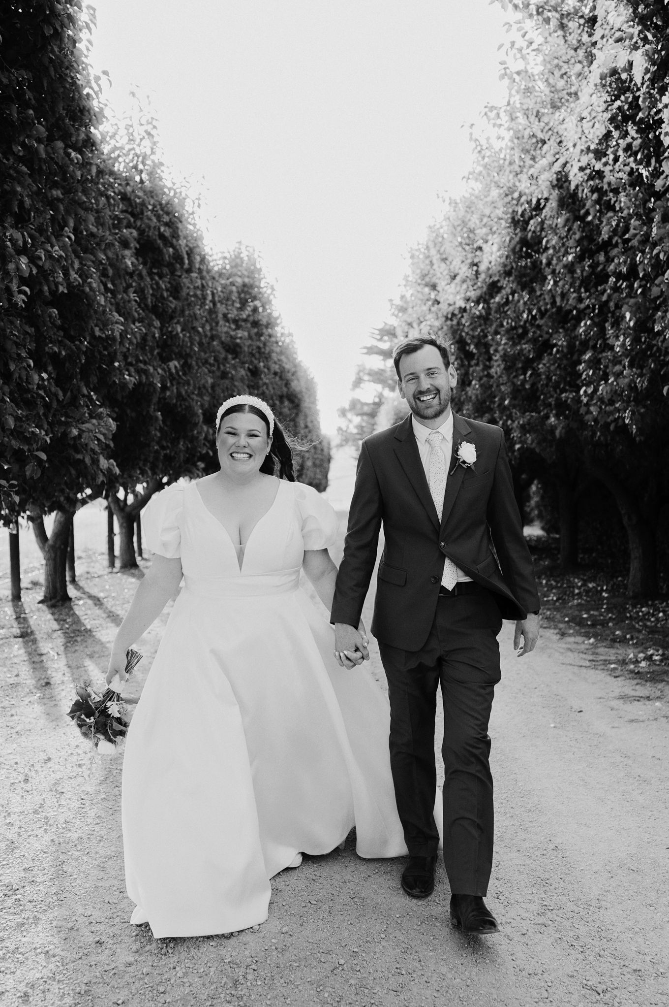 Black and white photo of newlywed couple walking and laughing together on a tree-lined path.