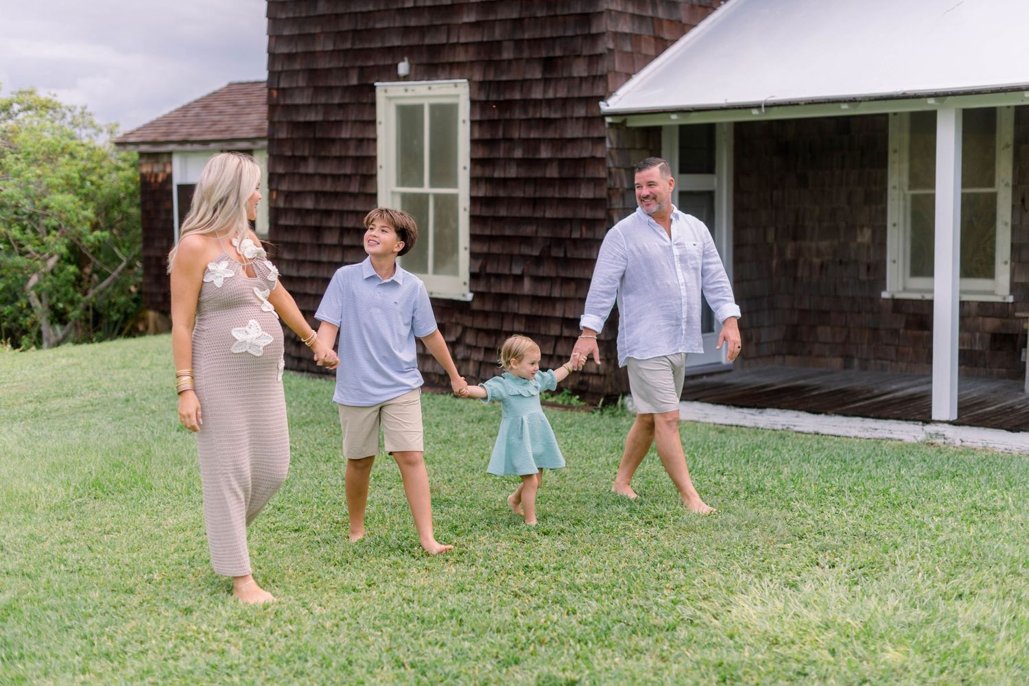 Family members holding hands and walking together on a grassy lawn outside a brown shingled house on a sunny day.