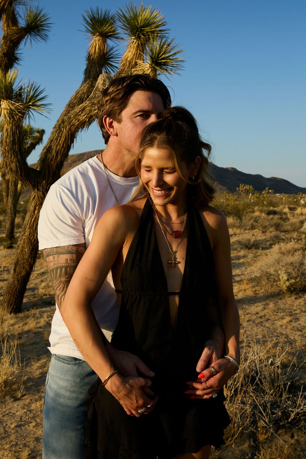 A couple shares a romantic moment at sunset in the desert with Joshua trees in the background.