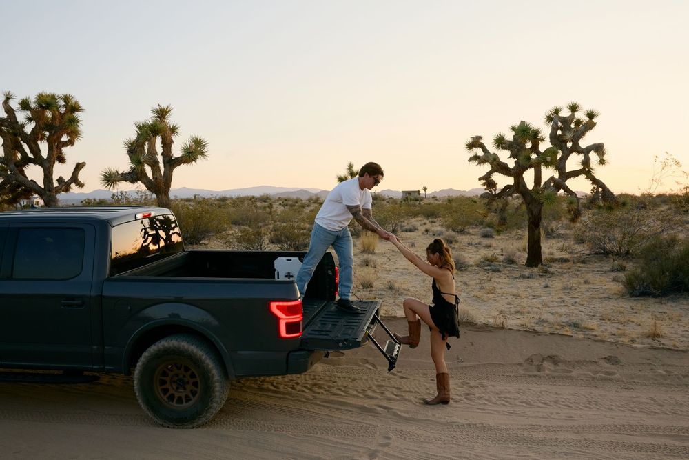 Two people load camping gear into a pickup truck at sunset in Joshua Tree National Park with desert landscape behind them.